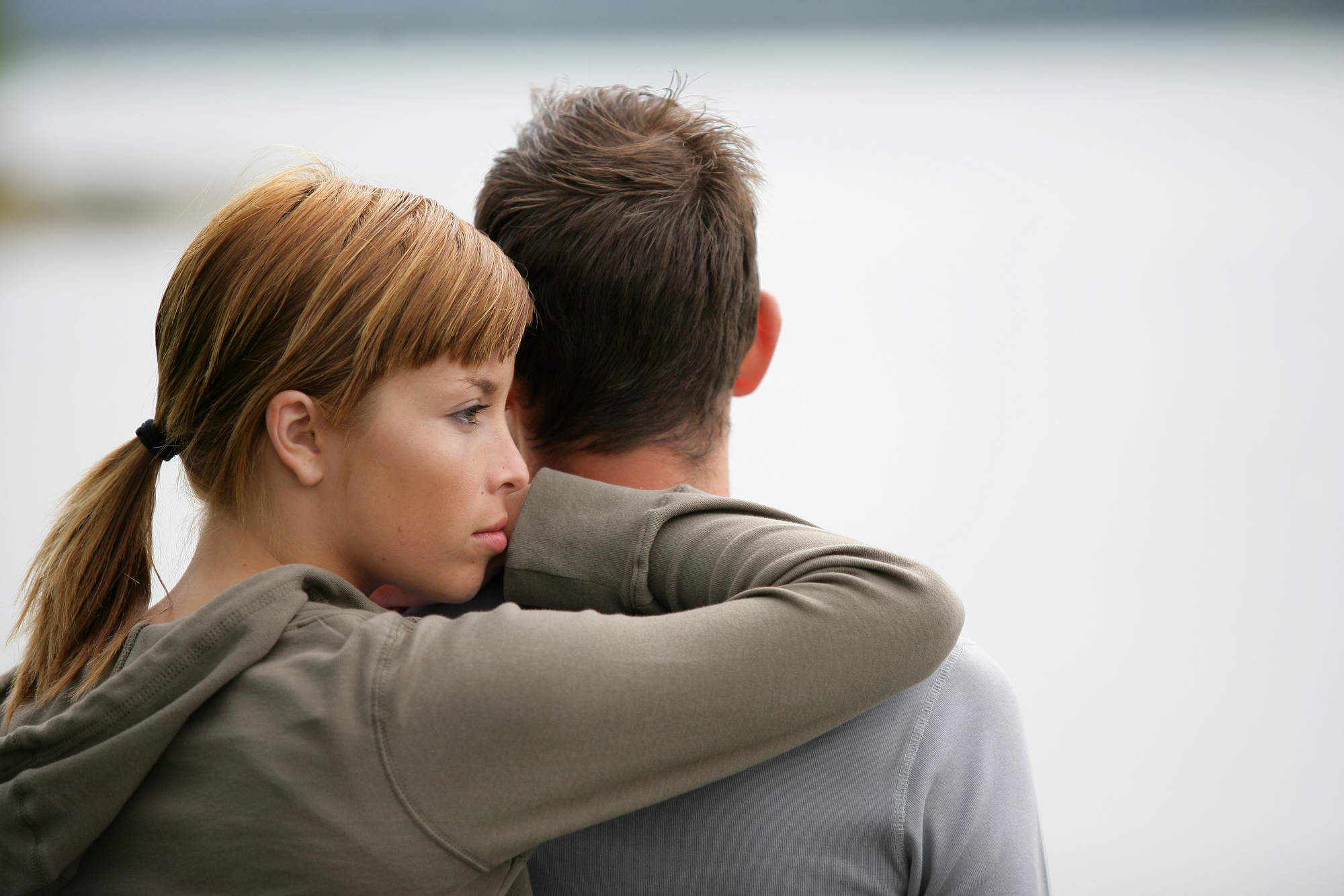 A woman with light brown hair rests her head on the shoulder of a man with short brown hair. They are standing close to each other, with the woman looking off into the distance. The background is blurry and appears to be an outdoor setting, possibly near water.