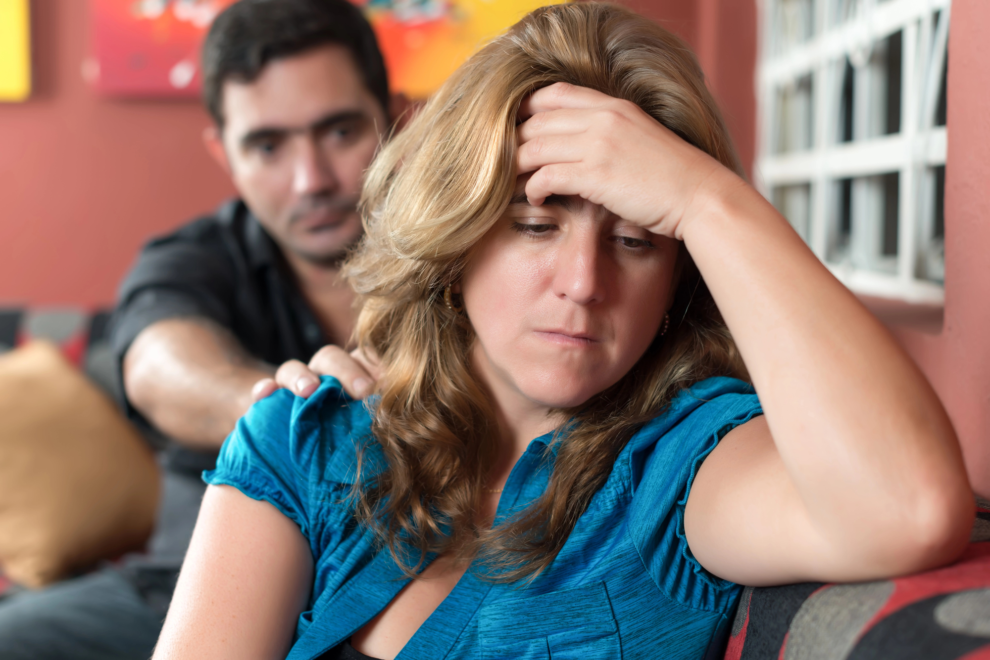 A woman with blonde hair, looking distressed, sits on a couch with her hand on her forehead. A man sits beside her, with a concerned expression, resting his hand on her shoulder in a comforting gesture. The background is softly blurred.