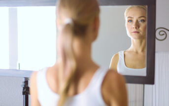 A woman with her hair tied back in a ponytail is seen from behind, looking at herself in a large mirror. She wears a white tank top and gazes at her reflection with a thoughtful expression. The background features a white wall and a decorative element.