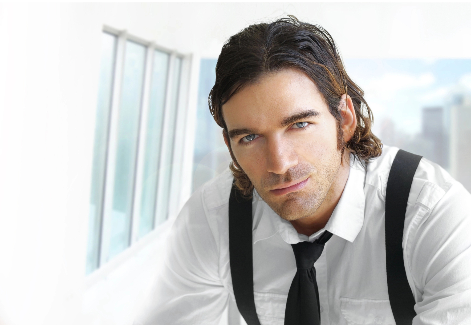 Man with medium-length dark hair and a beard wearing a white shirt, black tie, and black suspenders looks at the camera. The background is bright with windows and a blurred cityscape.