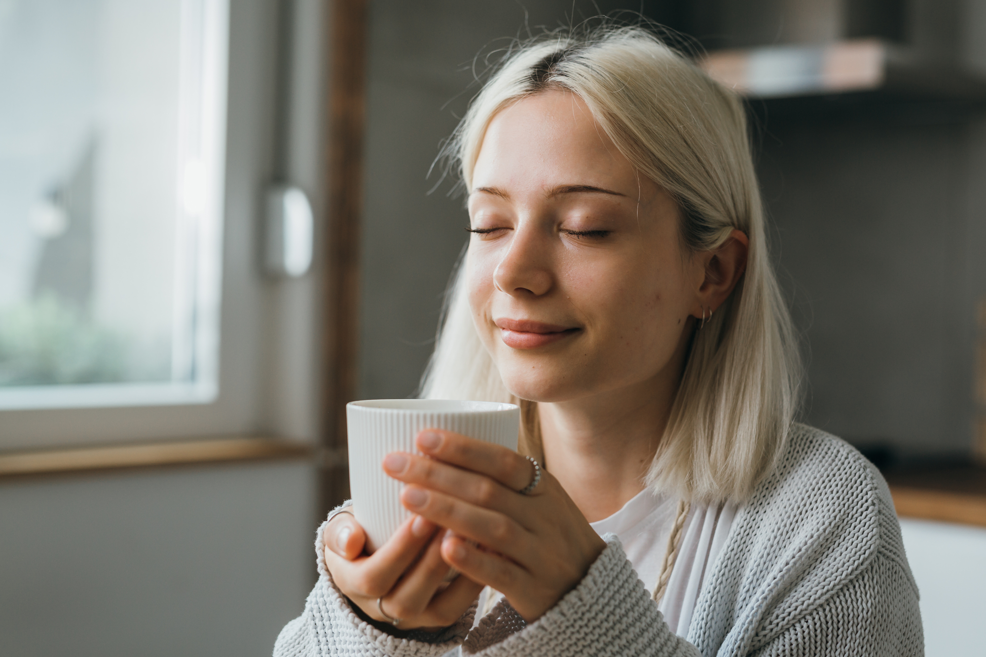 A woman with blonde hair holds a white mug with both hands, eyes closed, and a content smile on her face. She is wearing a light-colored cardigan and sits in a cozy indoor setting with a window in the background allowing natural light to gently illuminate her face.