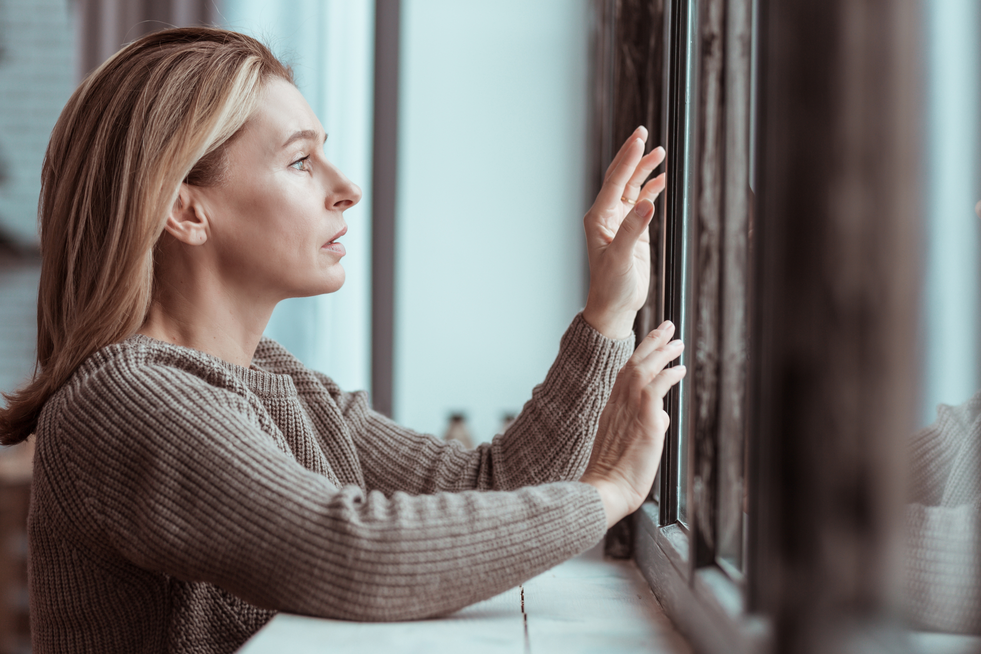 A woman with blonde hair, wearing a beige knit sweater, looks out a window with a pensive expression. One of her hands rests on the window frame while the other touches the glass. The atmosphere appears reflective and calm.