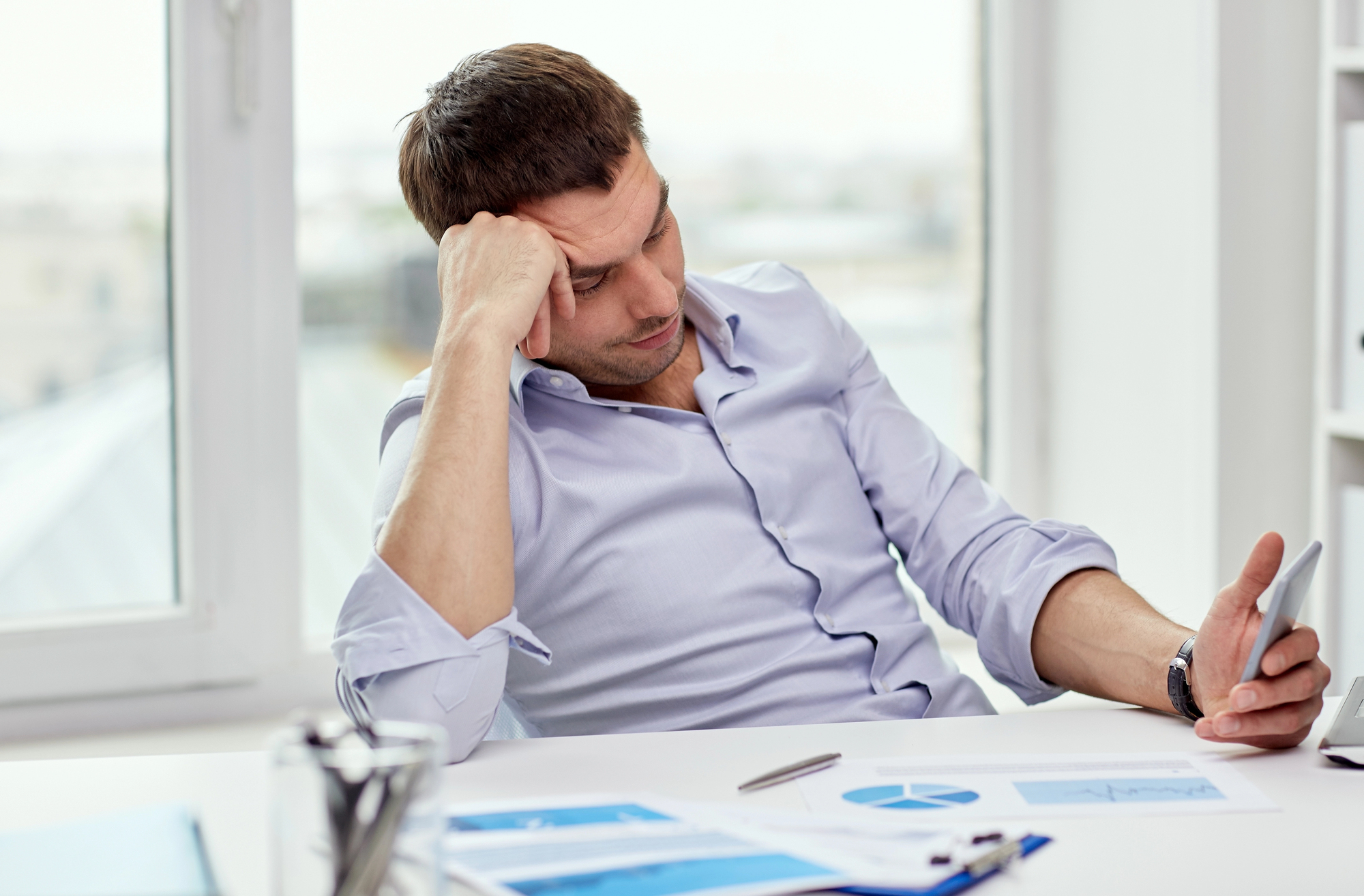 A man sitting at a desk, leaning his head on his hand and looking at his phone with a tired expression. He is surrounded by work-related items, including papers with charts and graphs, a pen, and office supplies. There is a window in the background.