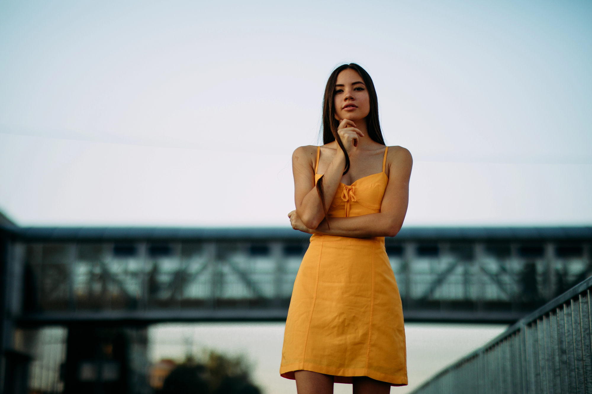 A woman with long hair, wearing a yellow sleeveless dress, stands pensively on an outdoor walkway. She has one hand on her chin and the other arm crossed, with a sky bridge in the blurred background. The image is taken during daytime.