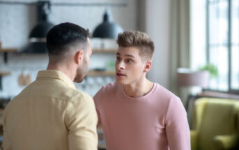 Two men are standing close to each other in a living room, engaged in a serious conversation. The man on the right, in a pink shirt, appears to be speaking intently to the man on the left, who is wearing a beige shirt. The background features a modern, well-lit space.