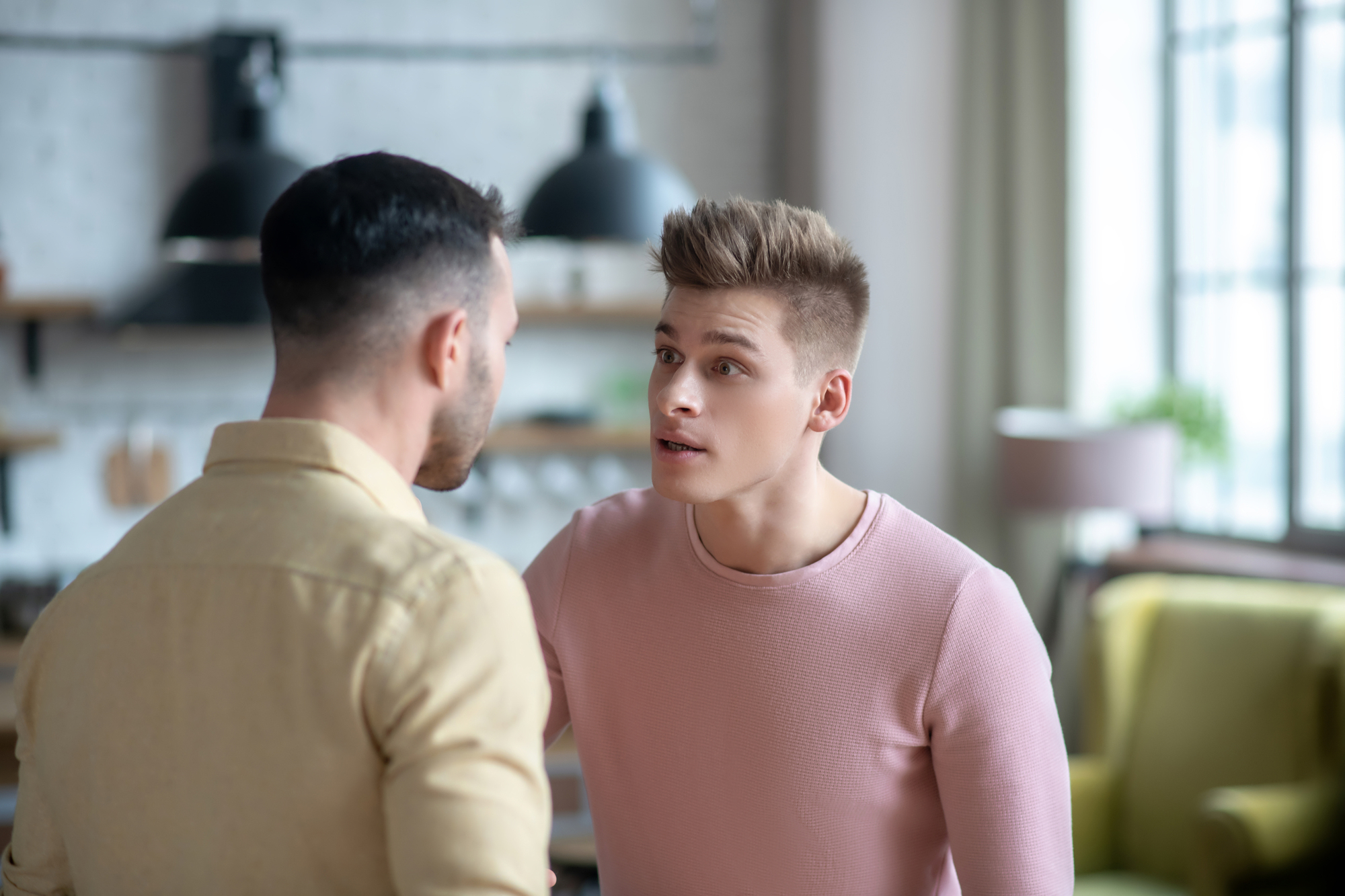 Two men are standing close to each other in a living room, engaged in a serious conversation. The man on the right, in a pink shirt, appears to be speaking intently to the man on the left, who is wearing a beige shirt. The background features a modern, well-lit space.