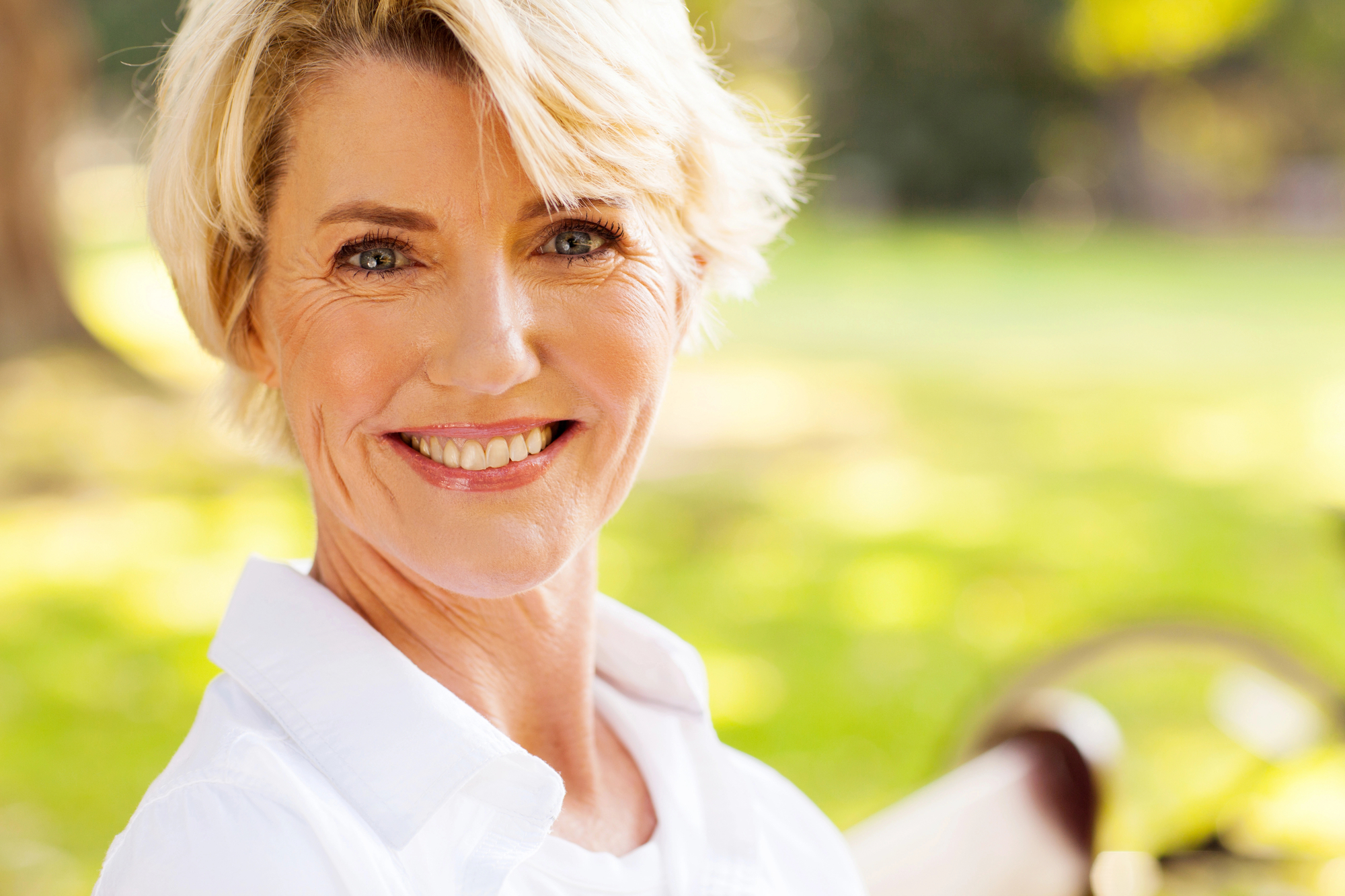 A smiling woman with short, blonde hair and wearing a white blouse is standing outdoors in a sunny park. The blurred background features green grass and trees, suggesting a serene and pleasant environment.