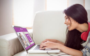 A woman with long dark hair lies on a sofa, smiling while typing on a laptop. She has red headphones around her neck. The laptop screen displays a website with a couple's photo and a login form. Sunlight filters through a window in the background.