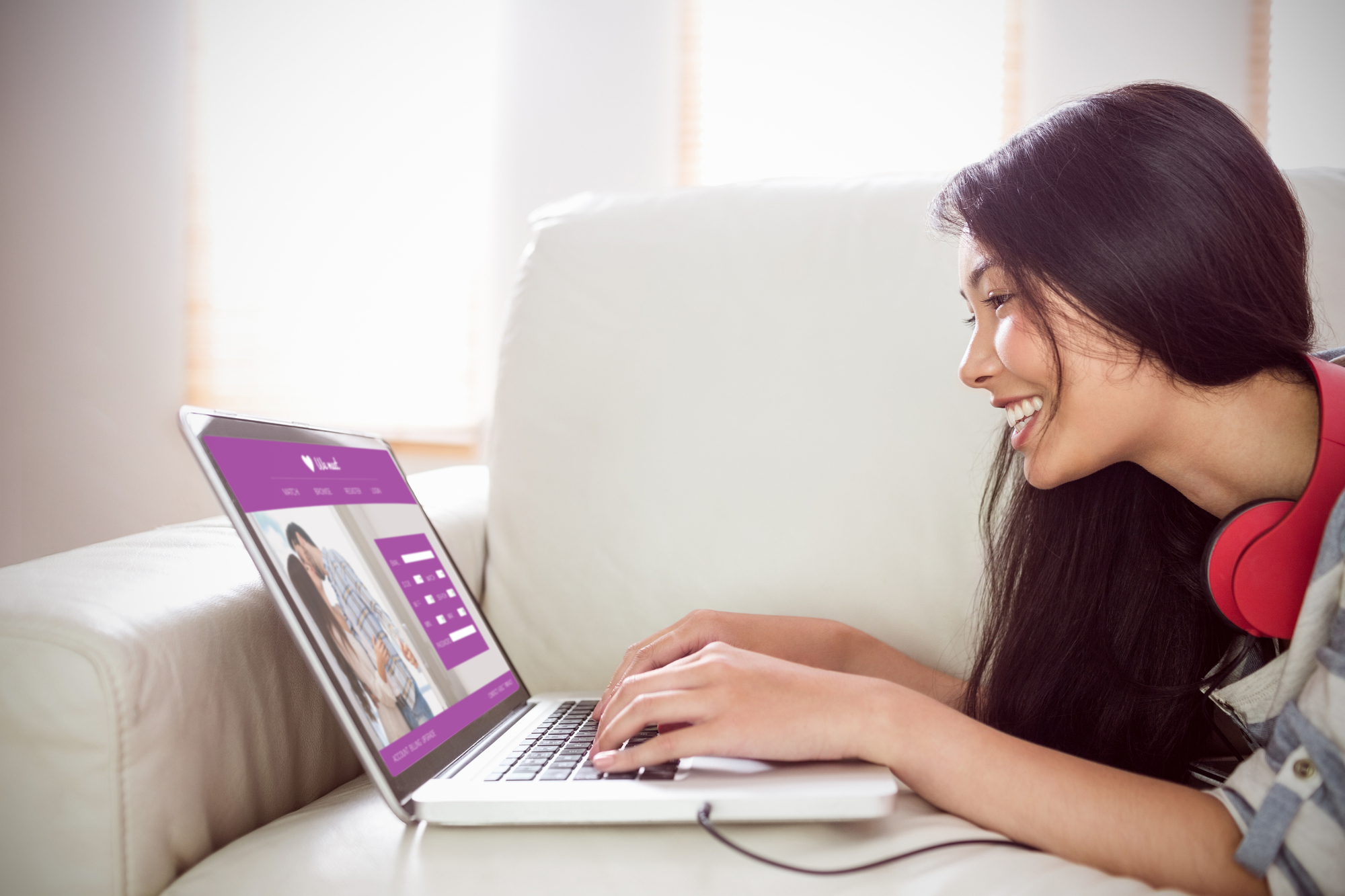 A woman with long dark hair lies on a sofa, smiling while typing on a laptop. She has red headphones around her neck. The laptop screen displays a website with a couple's photo and a login form. Sunlight filters through a window in the background.