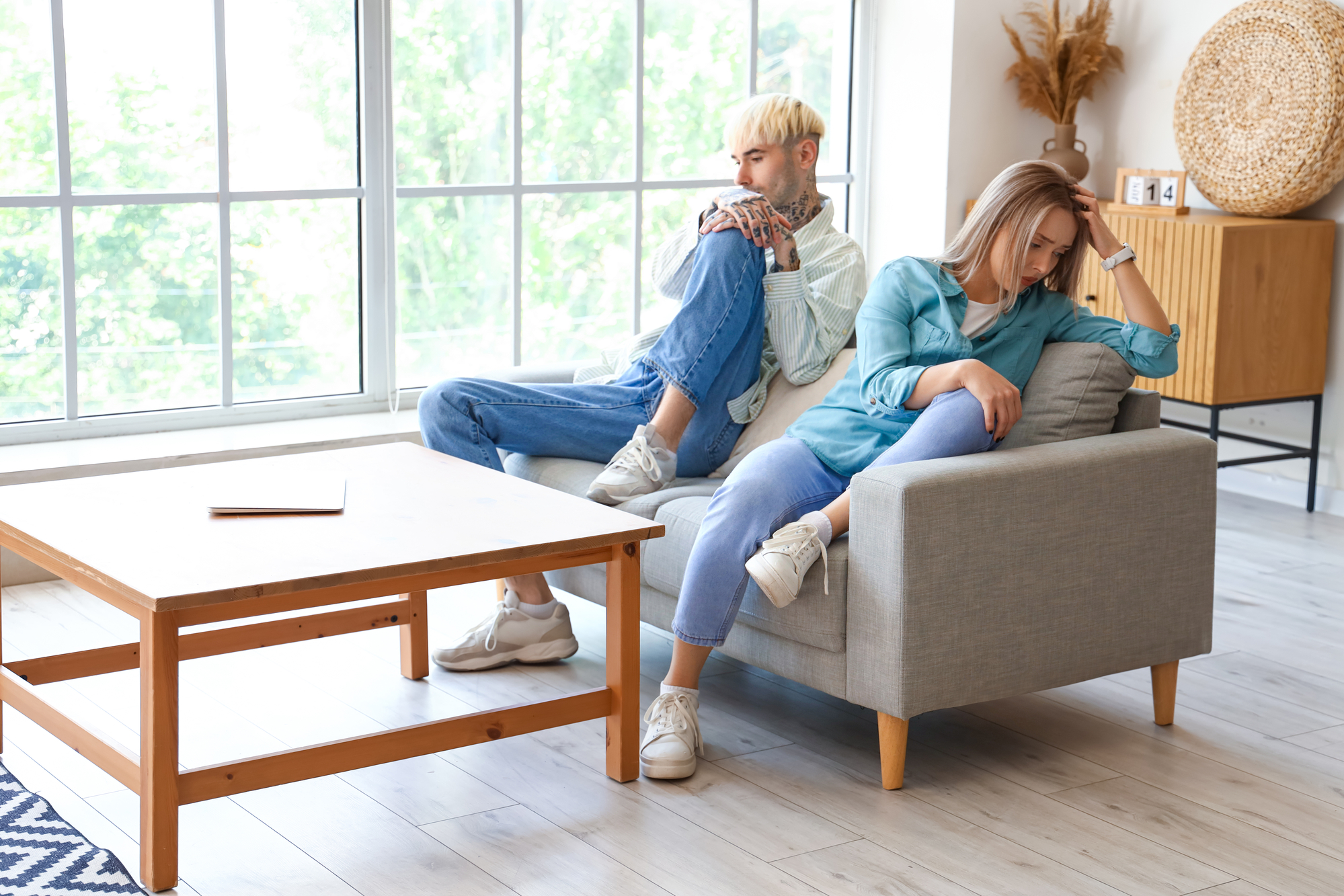 Two people, one with short blond hair and the other with long light hair, sit on a gray couch in a bright room with large windows. They appear upset, with one sitting cross-legged and the other leaning on one arm, looking away. A wooden coffee table is in front of them.