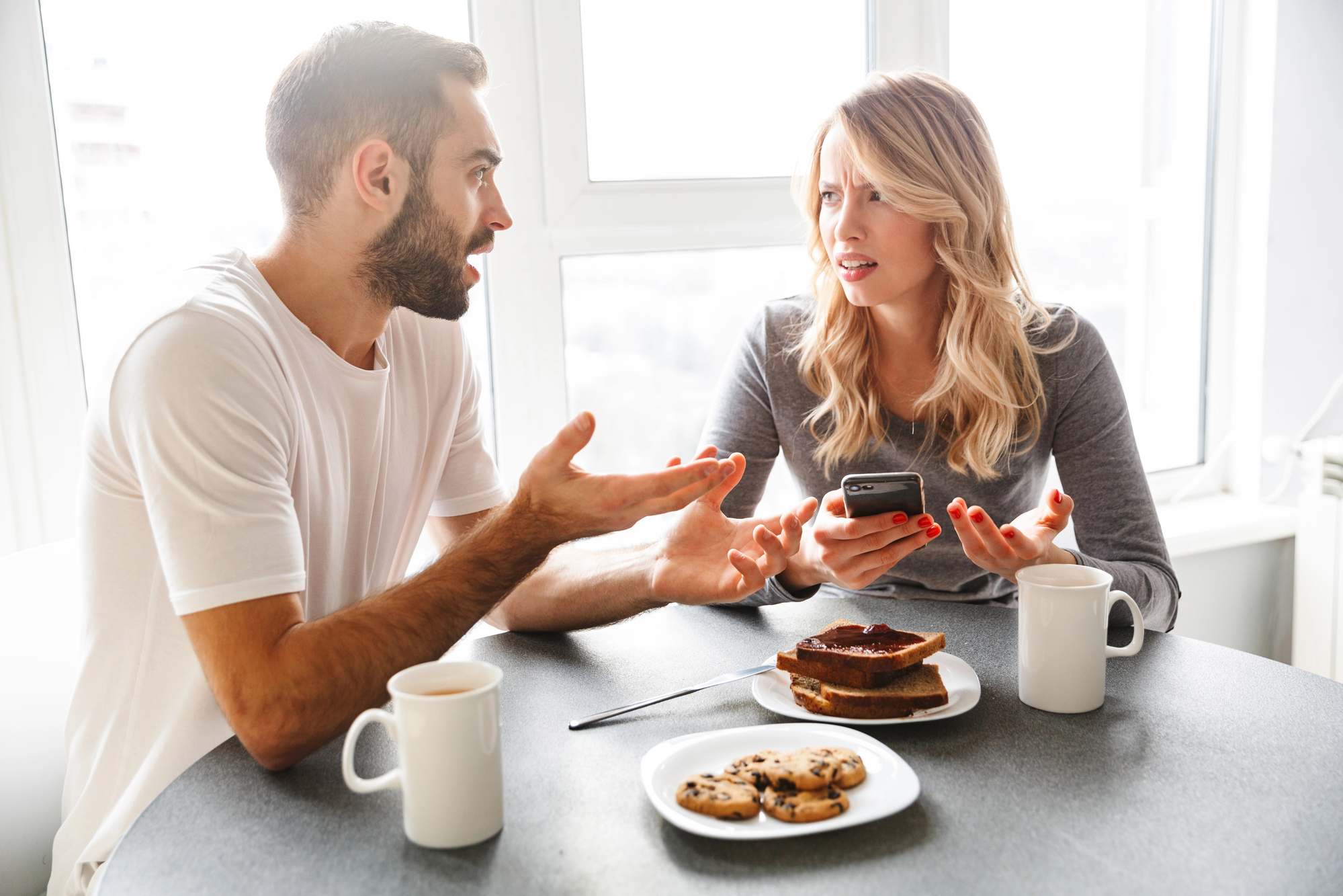 A man and woman are sitting at a table near a window, having a conversation. The man gestures with his hands, while the woman looks at him, holding a phone. On the table are two mugs, a plate with cookies, and a plate with toast and jam.