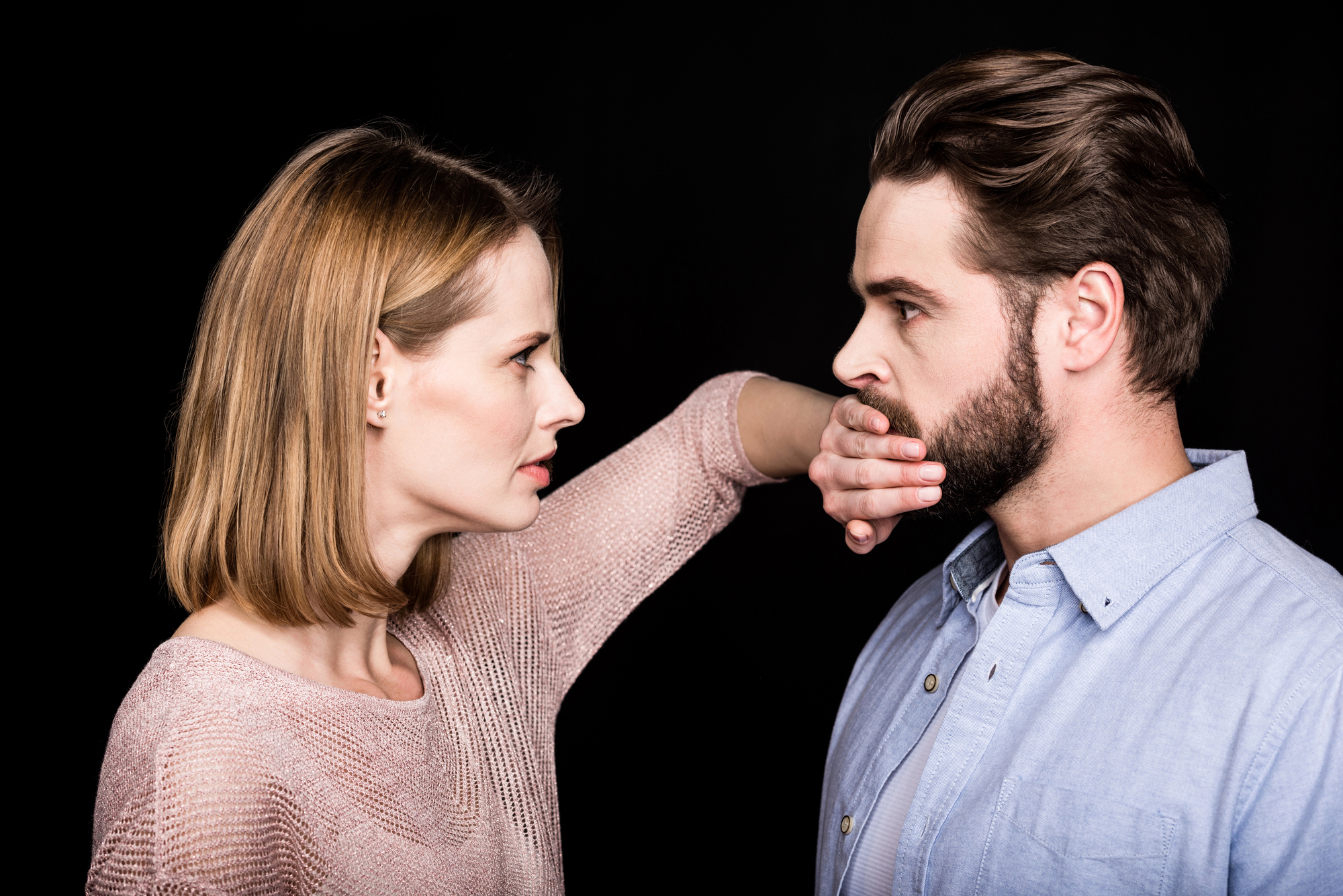 A woman with blonde hair is holding her hand over a bearded man's mouth as they face each other against a black background. The woman looks serious, while the man appears thoughtful. The woman is wearing a light pink sweater, and the man is wearing a light blue shirt.