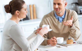 A man and woman sit at a table in a bright kitchen, enjoying coffee and pancakes. The man gestures with his hand while talking, and the woman holds a coffee cup, attentively listening. Both are casually dressed and appear engaged in conversation.