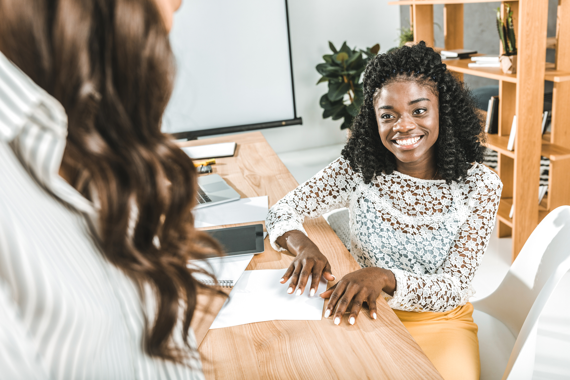 A woman with curly hair, wearing a white lacy top and yellow pants, smiles while sitting at a wooden desk. Another person, partially visible, stands with their back to the camera, engaging in conversation. The setting appears to be a modern office.