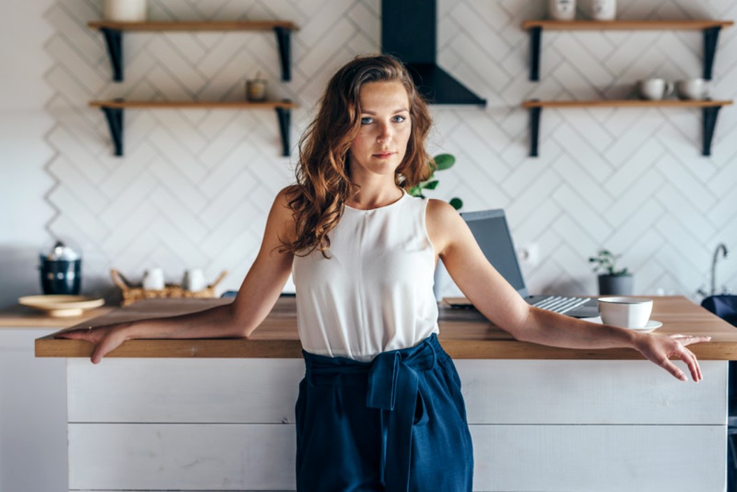 A woman with long, wavy hair is leaning against a kitchen island, dressed in a white sleeveless top and blue pants. She stands in a modern kitchen with shelves, various kitchen items, and a cup of coffee on the counter. A laptop is open in the background.