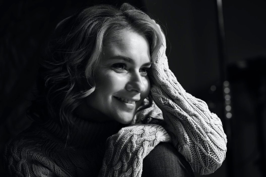 Black and white photo of a person smiling, resting their head on their right hand. They have wavy hair, wear a knitted sweater, and look off to the side. The lighting highlights their face, creating a warm and intimate atmosphere.