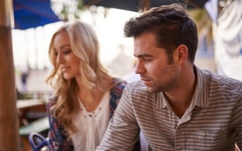 A man and a woman sit together at an outdoor cafe. The man has short dark hair and a striped shirt, looking contemplatively to the side. The woman has long blonde hair and is slightly blurred, wearing a patterned cardigan over a white top.