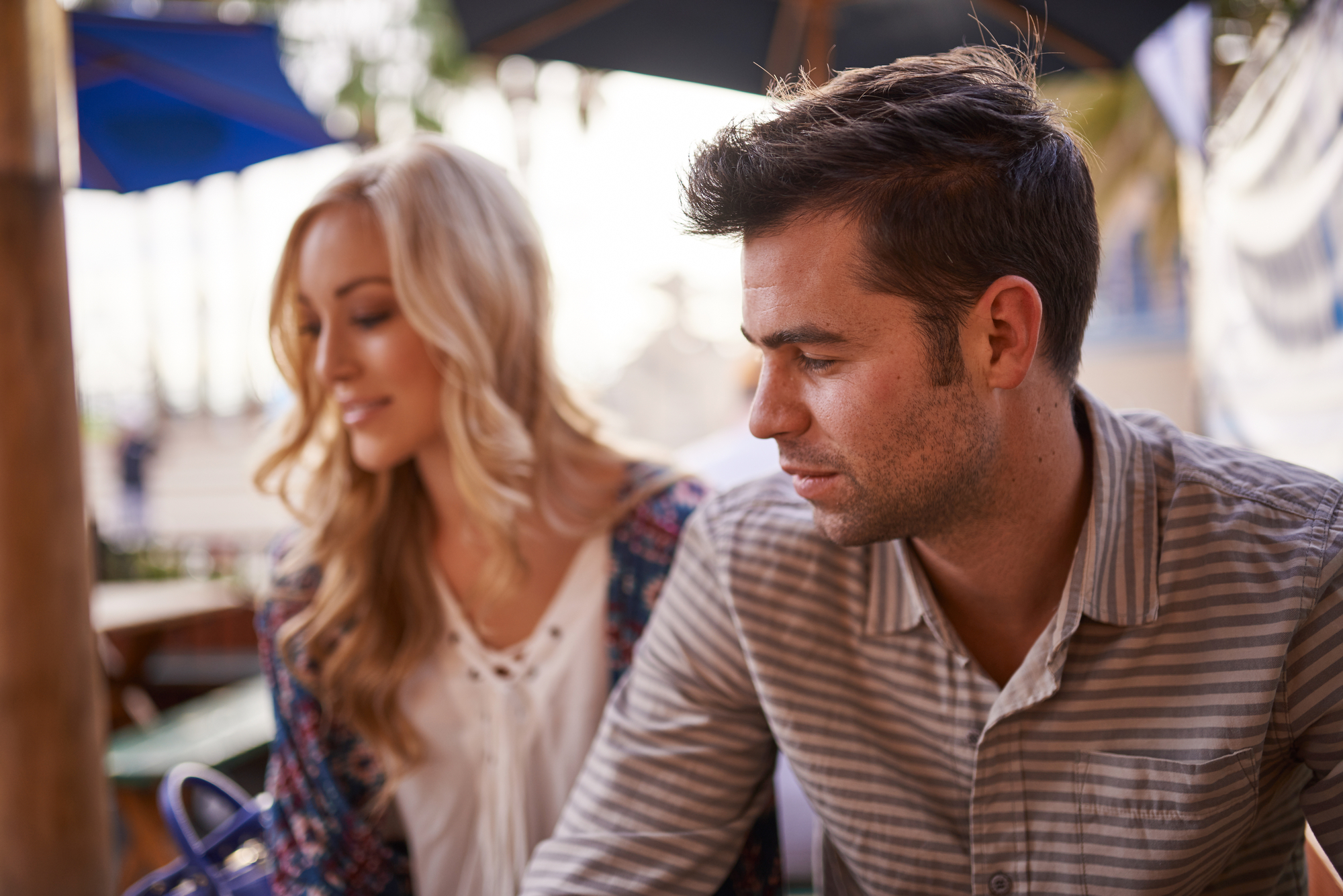 A man and a woman sit together at an outdoor cafe. The man has short dark hair and a striped shirt, looking contemplatively to the side. The woman has long blonde hair and is slightly blurred, wearing a patterned cardigan over a white top.