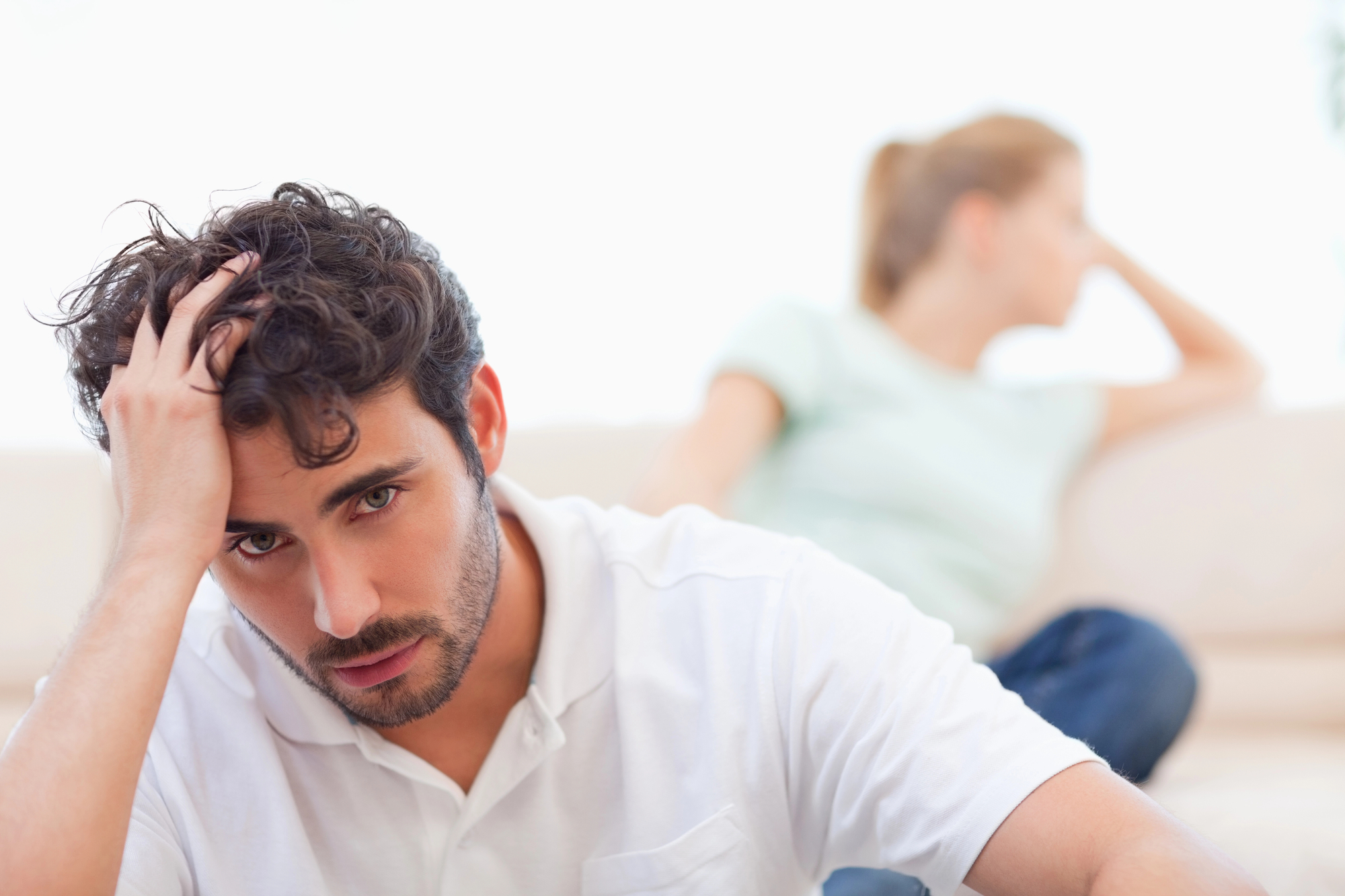 A man in a white shirt sits in the foreground with a distressed expression, holding his head with one hand. In the background, a woman in a light green shirt sits turned away, gazing into the distance. Both appear to be in a tense or thoughtful mood.