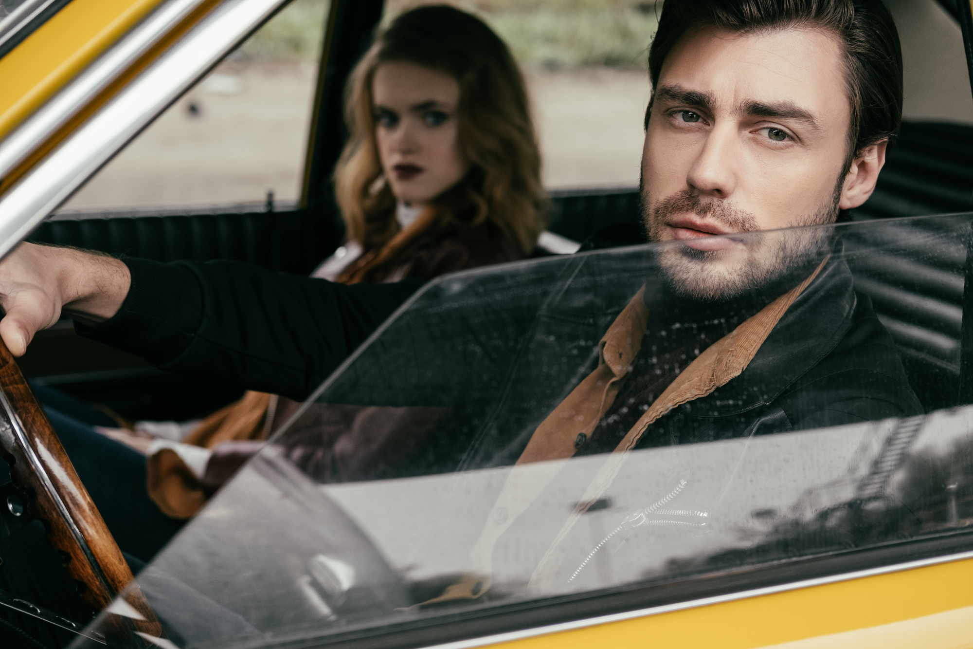 A man with a serious expression sits behind the wheel of a yellow car, with his hand on the steering wheel. A woman with long hair and wearing dark lipstick sits in the passenger seat, looking straight ahead. The car window is partly rolled down.