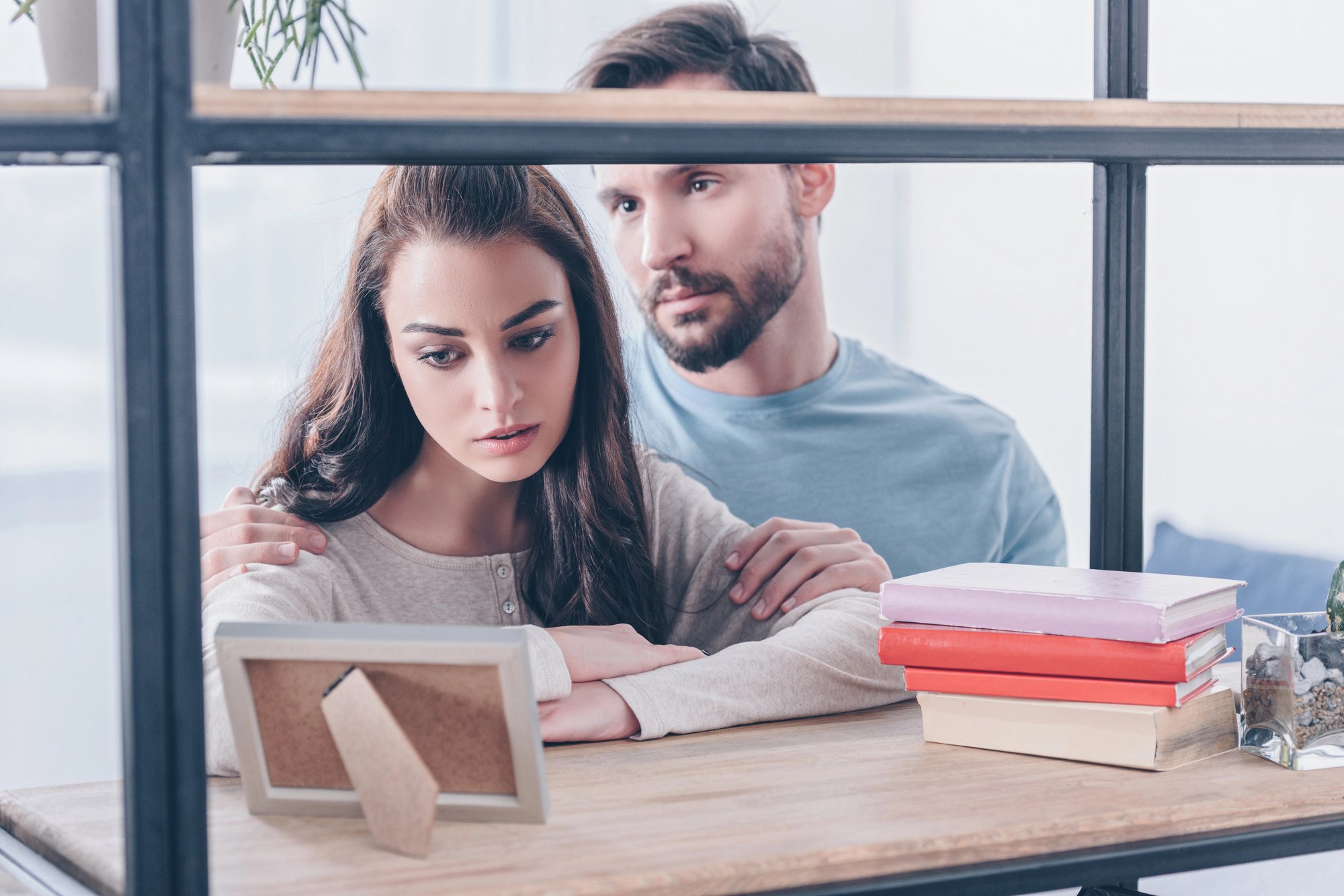 A woman and man are sitting together by a window, looking at a picture frame on a wooden table. The woman looks pensive with the man's arm gently around her shoulders. A stack of books sits on the table beside the frame.