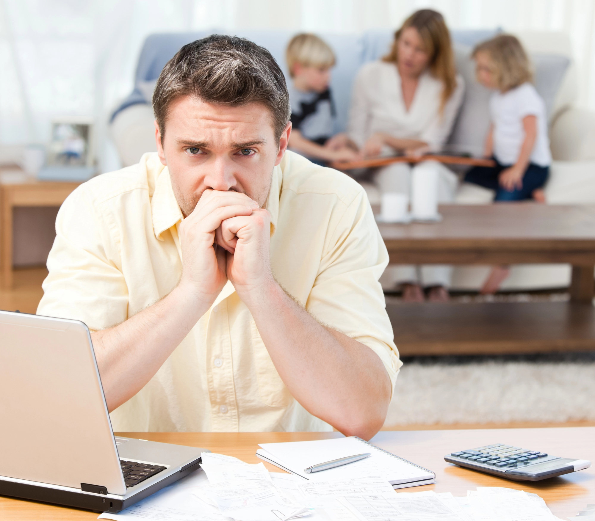 A worried man sits at a desk with a laptop, notebook, and calculator, surrounded by papers. In the background, a woman and two children are sitting on a couch, engaged in an activity. The scene suggests stress or concern about financial matters.