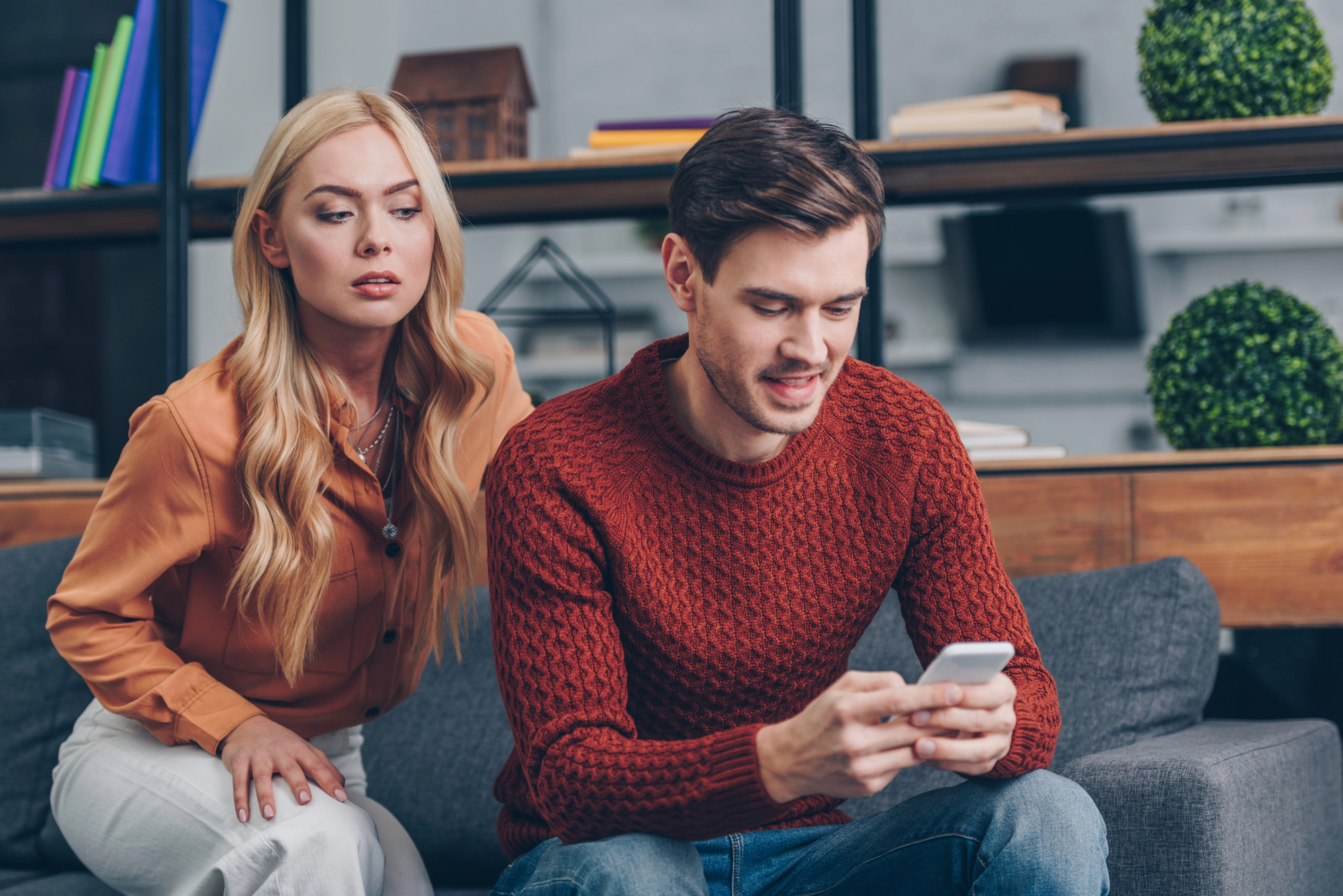 A man in a red sweater sits on a gray couch, engrossed in his smartphone. A woman with long blonde hair and wearing an orange blouse leans over from behind, seemingly curious about what he is looking at. Shelves with books and decor are in the background.