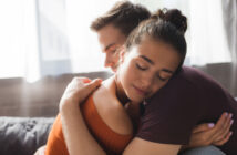 Two people are embracing each other lovingly in a bright room. The person facing the camera has her eyes closed, wearing an orange top. The person facing away has their eyes closed, with their head resting near her shoulder. Both appear calm and content.