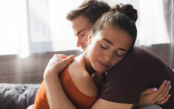 Two people are embracing each other lovingly in a bright room. The person facing the camera has her eyes closed, wearing an orange top. The person facing away has their eyes closed, with their head resting near her shoulder. Both appear calm and content.