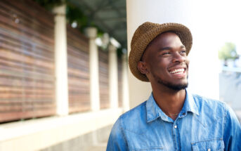 A man wearing a brown straw hat and a blue denim shirt smiles while standing outdoors. The background features a wooden fence and a covered walkway.