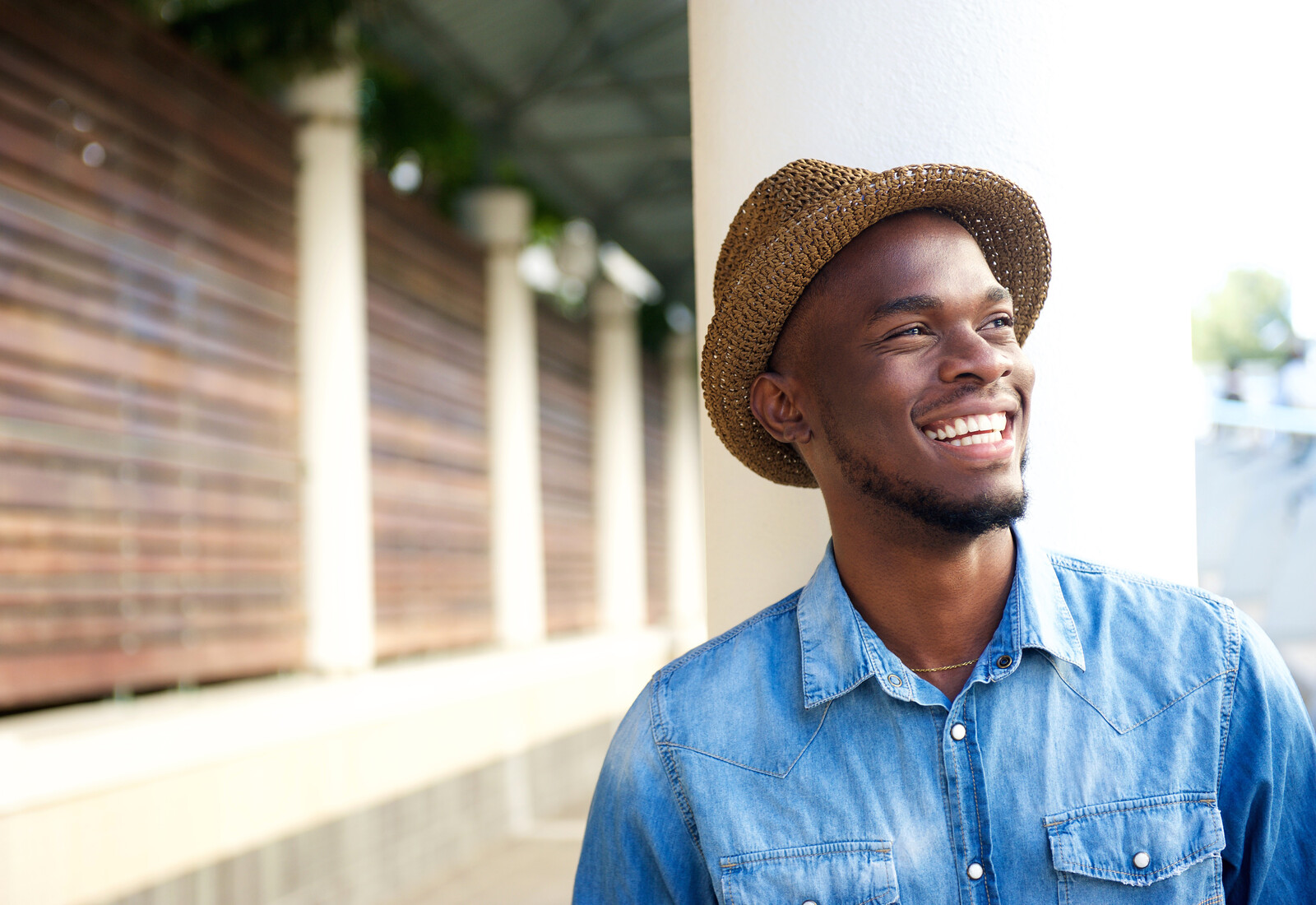 A man wearing a brown straw hat and a blue denim shirt smiles while standing outdoors. The background features a wooden fence and a covered walkway.