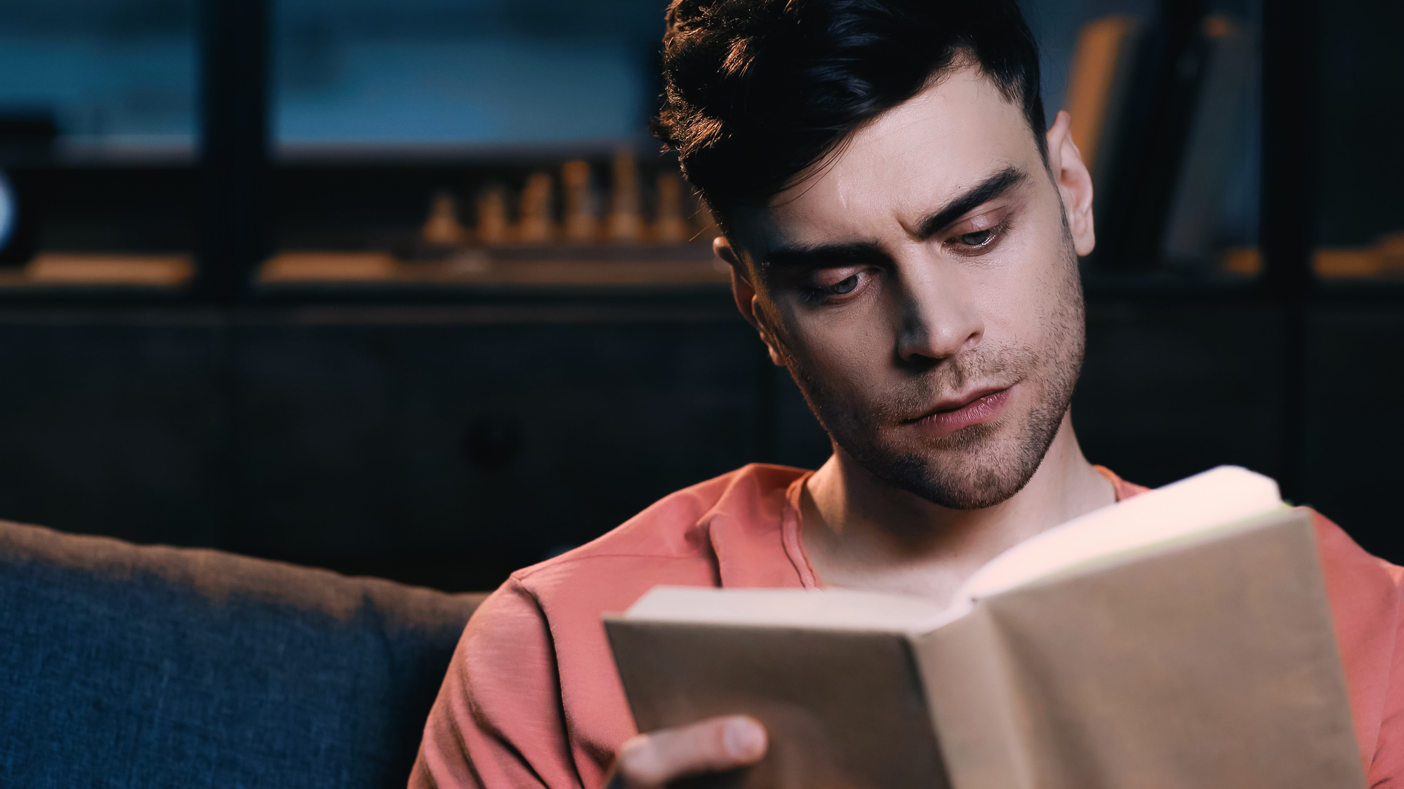 A man with dark hair, wearing a coral-colored shirt, sits on a couch and reads an open book. The background is dimly lit with blurred shelves holding books and a chess set. The man appears focused on his reading.