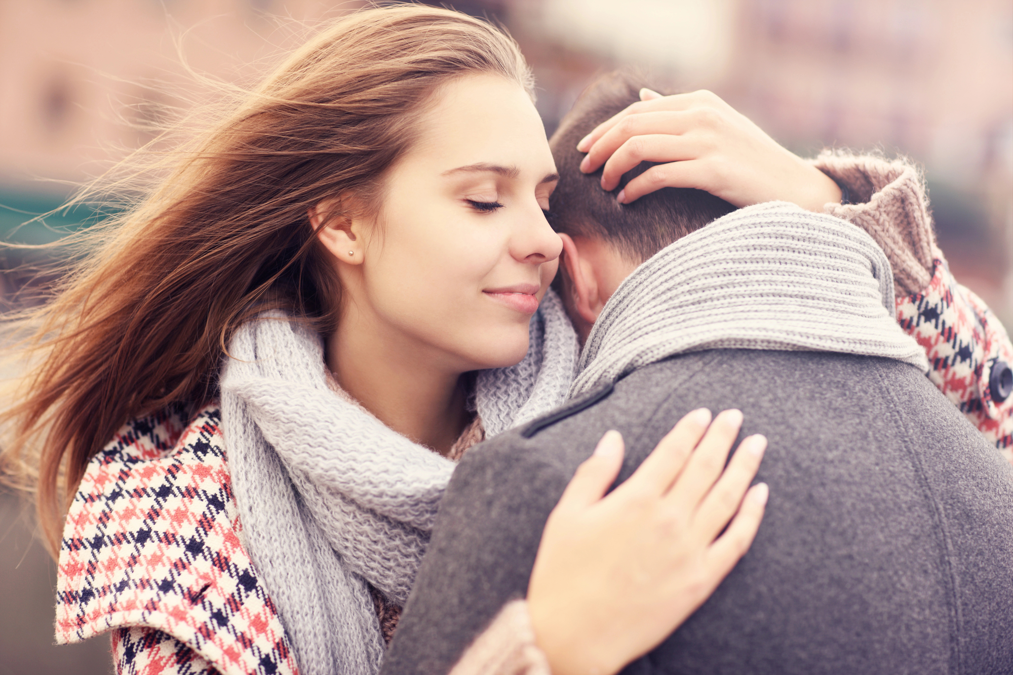 A woman with long blonde hair and a gray scarf closes her eyes and smiles while hugging a man dressed in a gray coat and scarf. She gently places her hand on the back of his head. They are outdoors on a chilly day.