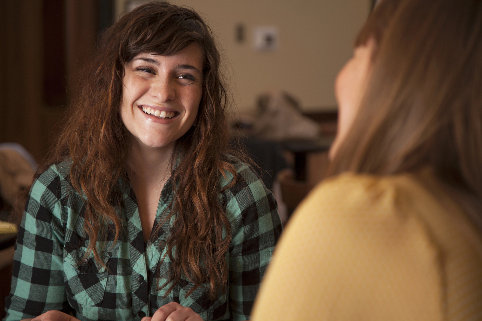 A woman with long curly hair, wearing a green plaid shirt, sits and smiles at another woman in the foreground who is blurred. They appear to be having a conversation in a cozy indoor setting.