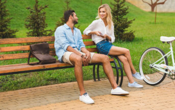 A man and a woman are sitting on a park bench. The man, wearing a light blue shirt and shorts, is looking at the woman. The woman, in a white blouse and denim shorts, is smiling and holding a smartphone. A white bicycle is parked next to the bench.