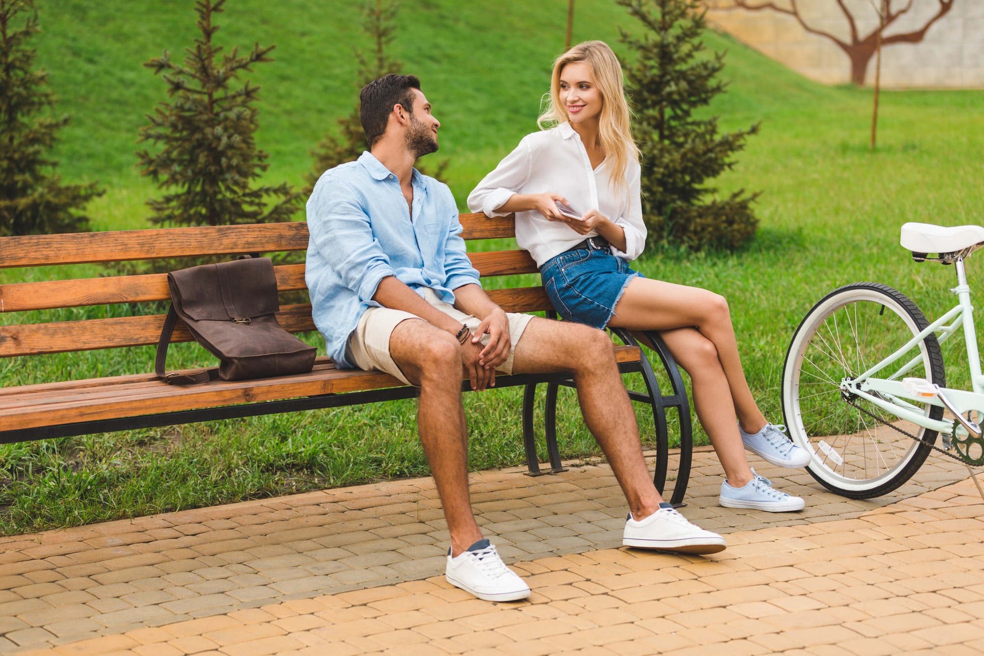 A man and a woman are sitting on a park bench. The man, wearing a light blue shirt and shorts, is looking at the woman. The woman, in a white blouse and denim shorts, is smiling and holding a smartphone. A white bicycle is parked next to the bench.