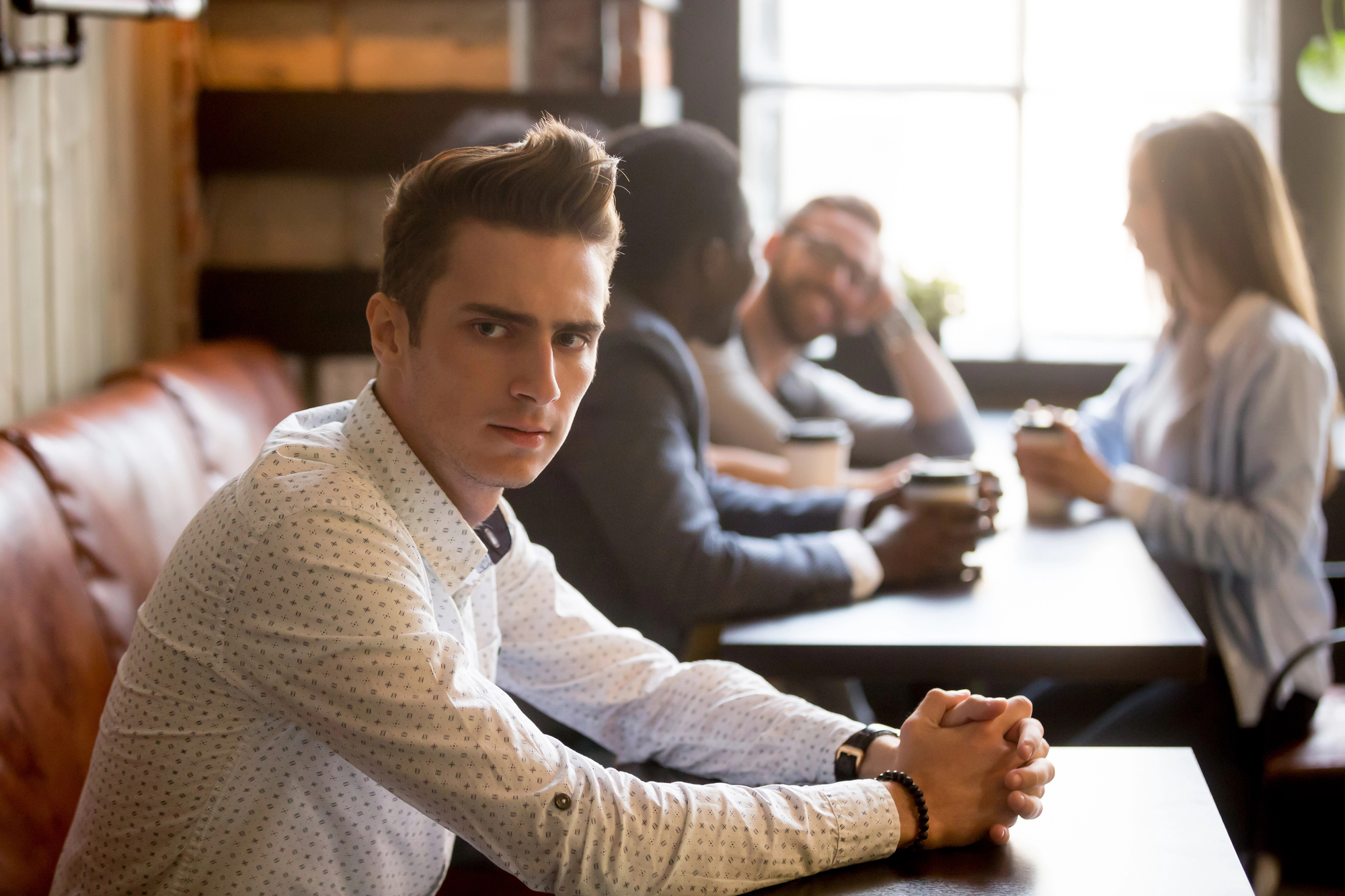 A man in a white shirt sits at a table in a café, looking directly at the camera with a serious expression. In the background, three other people are engaged in conversation, smiling and holding coffee cups. The café has a warm, cozy ambiance.