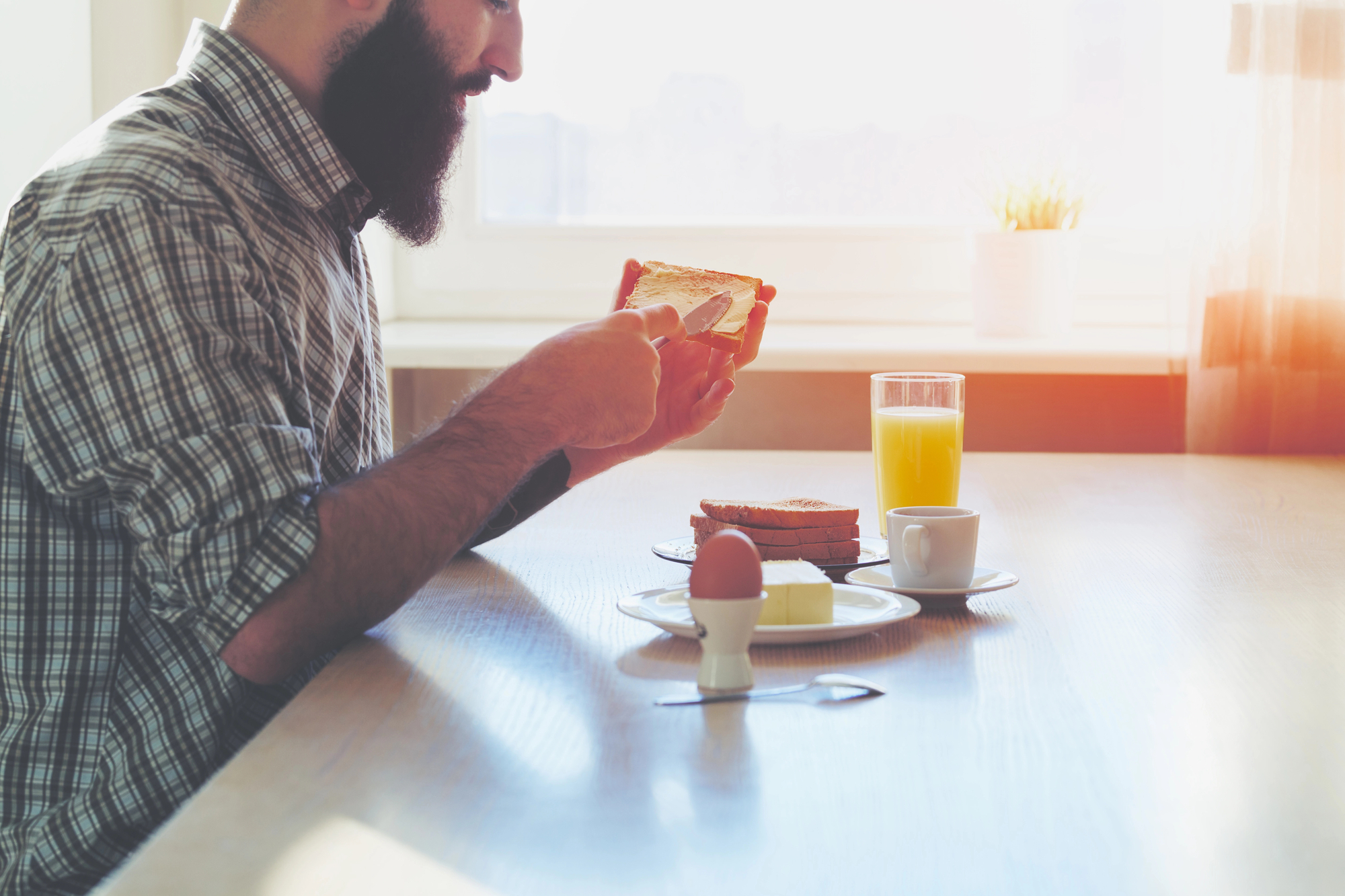 A bearded man in a plaid shirt spreads butter on a slice of toast at a sunlit breakfast table. The table also has stacked toast, a soft-boiled egg, a butter dish, a cup of coffee, and a glass of orange juice. The room is bright with sunlight coming through a window.