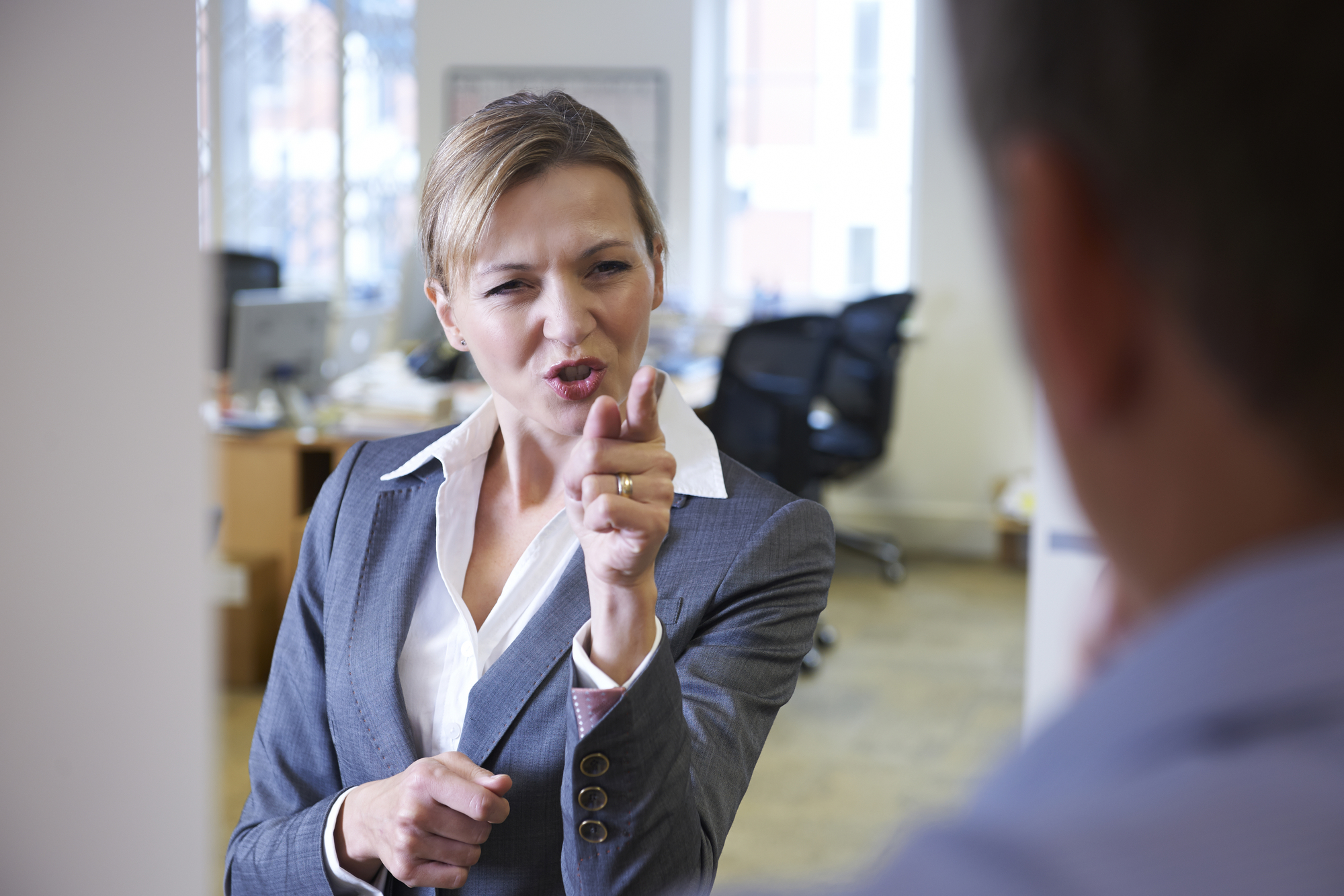 A woman in a business suit stands in an office, pointing her finger and appearing agitated while speaking to a man whose back is turned to the camera. Various office furniture and equipment, including desks and chairs, are visible in the background.