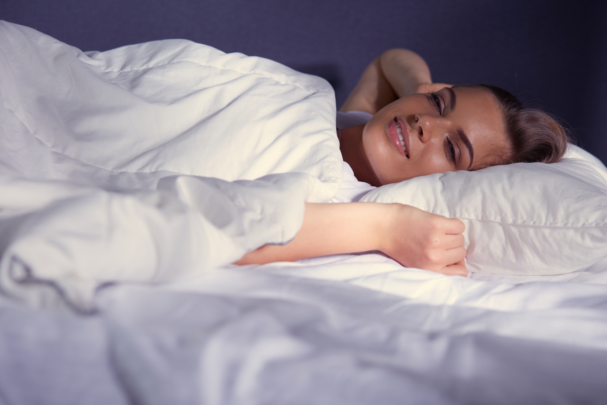 A person with blonde hair lies in bed, wrapped in a white blanket and resting their head on a white pillow. They appear to be smiling with eyes closed, in a peaceful and relaxed state. The background is dark.