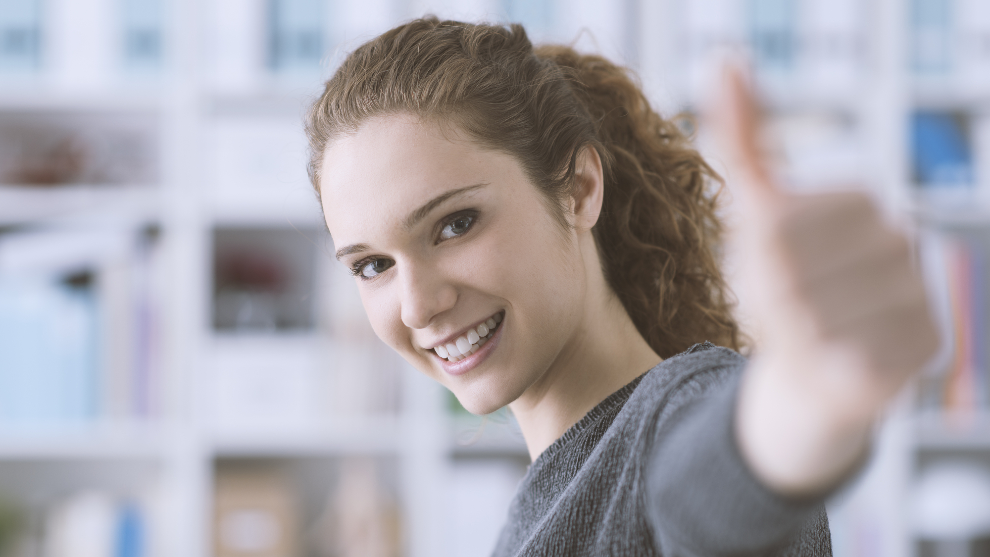 A woman with curly brown hair is smiling and giving a thumbs-up gesture. She is wearing a gray sweater and is standing in front of a blurry background filled with shelves.