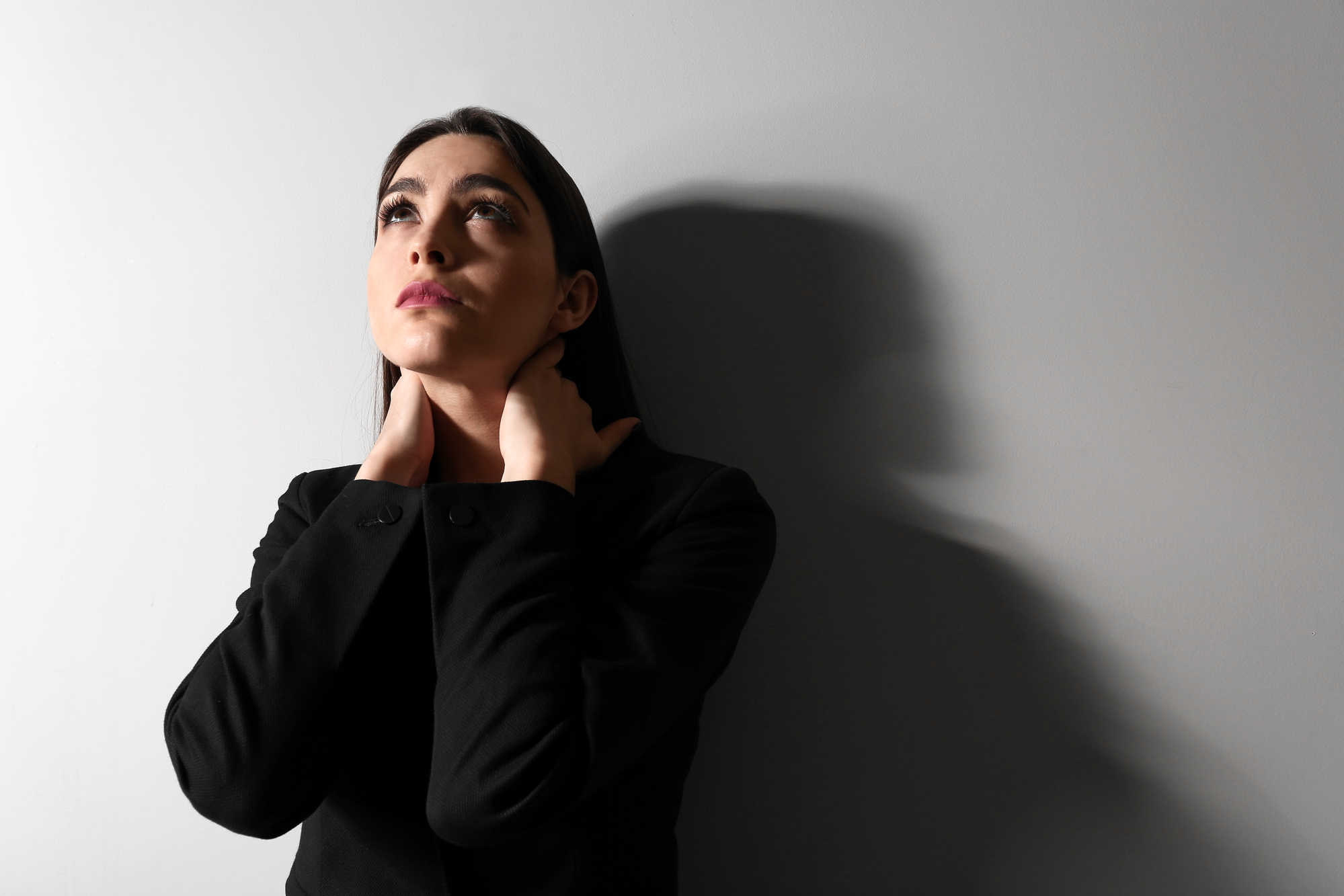 A woman with long dark hair and wearing a black outfit stands against a gray wall. She looks slightly upward with a contemplative expression, her hands gently touching her neck. The lighting creates a shadow of her on the wall.