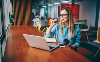 A woman with long blonde hair and glasses, dressed in a denim shirt, sits at a wooden table with a laptop, smartphone, and notebook in front of her. She is holding a pen near her mouth and a coffee cup in her other hand, looking thoughtfully out the window.