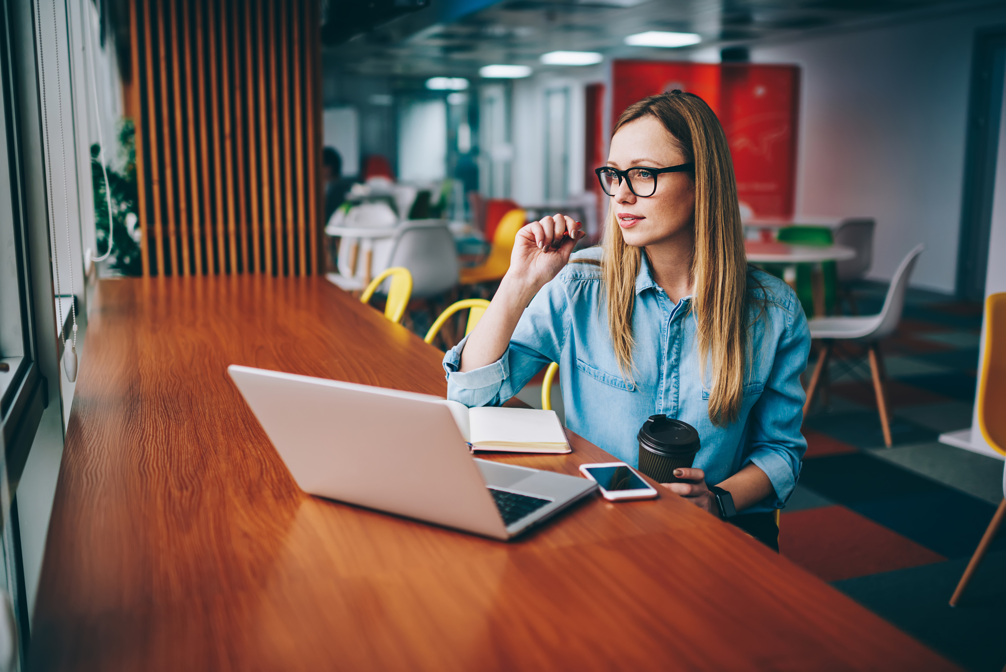 A woman with long blonde hair and glasses, dressed in a denim shirt, sits at a wooden table with a laptop, smartphone, and notebook in front of her. She is holding a pen near her mouth and a coffee cup in her other hand, looking thoughtfully out the window.