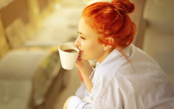 A person with bright red hair is sitting outdoors, sipping coffee from a white mug, and looking into the distance. They are wearing a light-colored, checkered shirt and have their hair tied up in a bun. The background is slightly blurred and shows a street with cars.