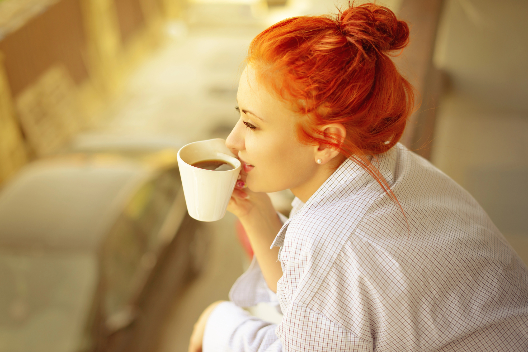 A person with bright red hair is sitting outdoors, sipping coffee from a white mug, and looking into the distance. They are wearing a light-colored, checkered shirt and have their hair tied up in a bun. The background is slightly blurred and shows a street with cars.