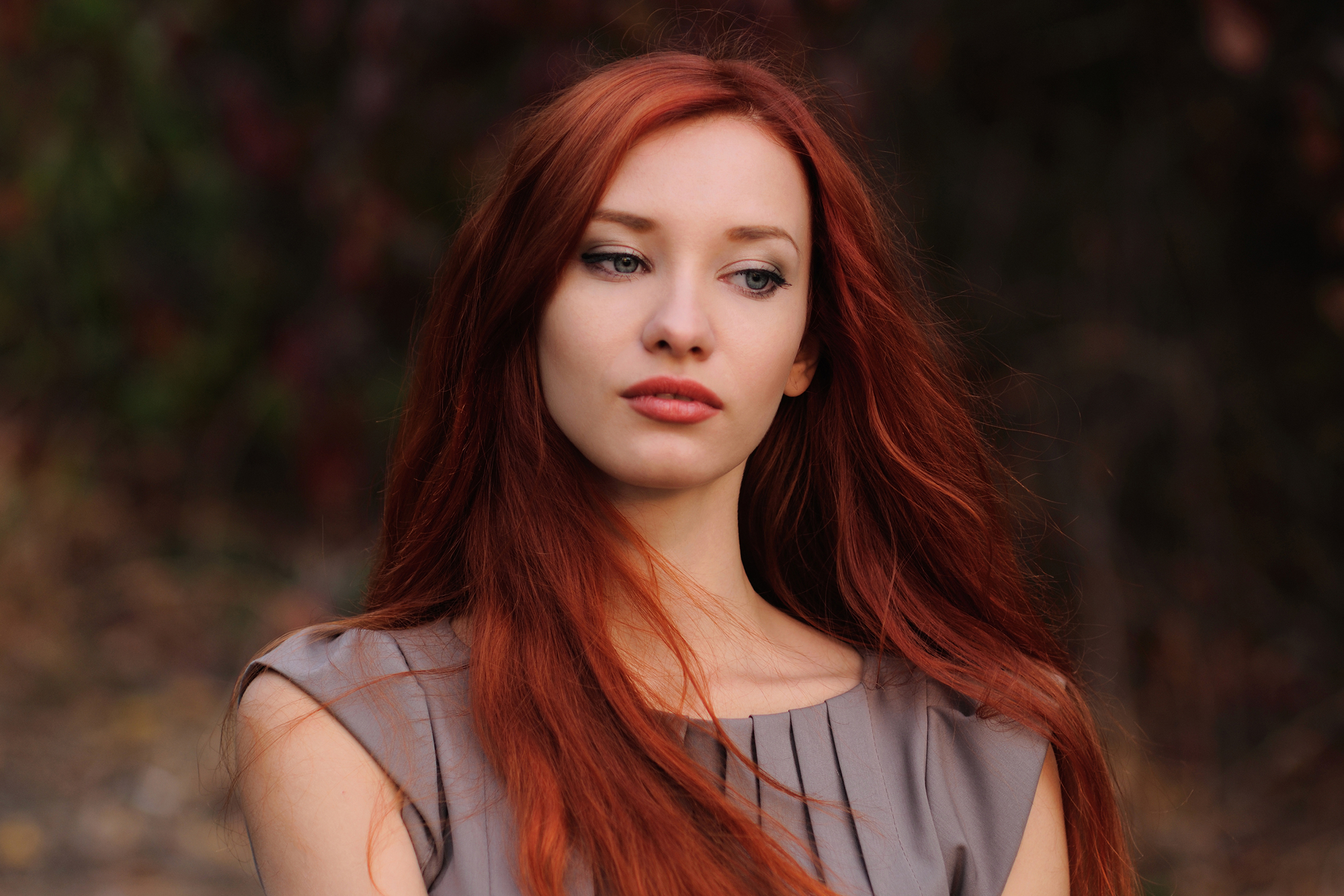 A young woman with long, flowing red hair stands outdoors against a blurred background of dark foliage. She wears a light brown sleeveless top and looks off to the side with a thoughtful expression.