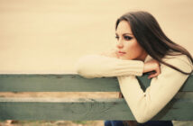 A woman with long brown hair and a cream sweater rests her arms on the back of a weathered wooden bench, gazing thoughtfully into the distance. The background is blurred, suggesting an outdoor setting on a calm day.