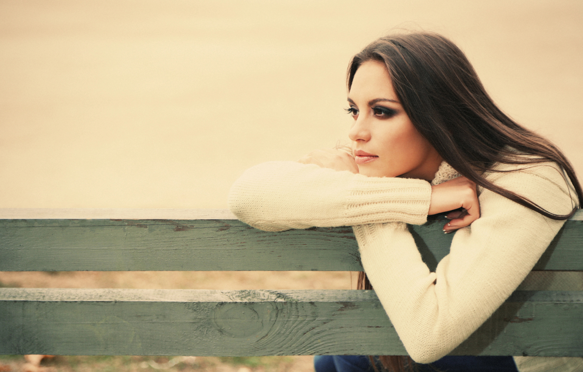 A woman with long brown hair and a cream sweater rests her arms on the back of a weathered wooden bench, gazing thoughtfully into the distance. The background is blurred, suggesting an outdoor setting on a calm day.
