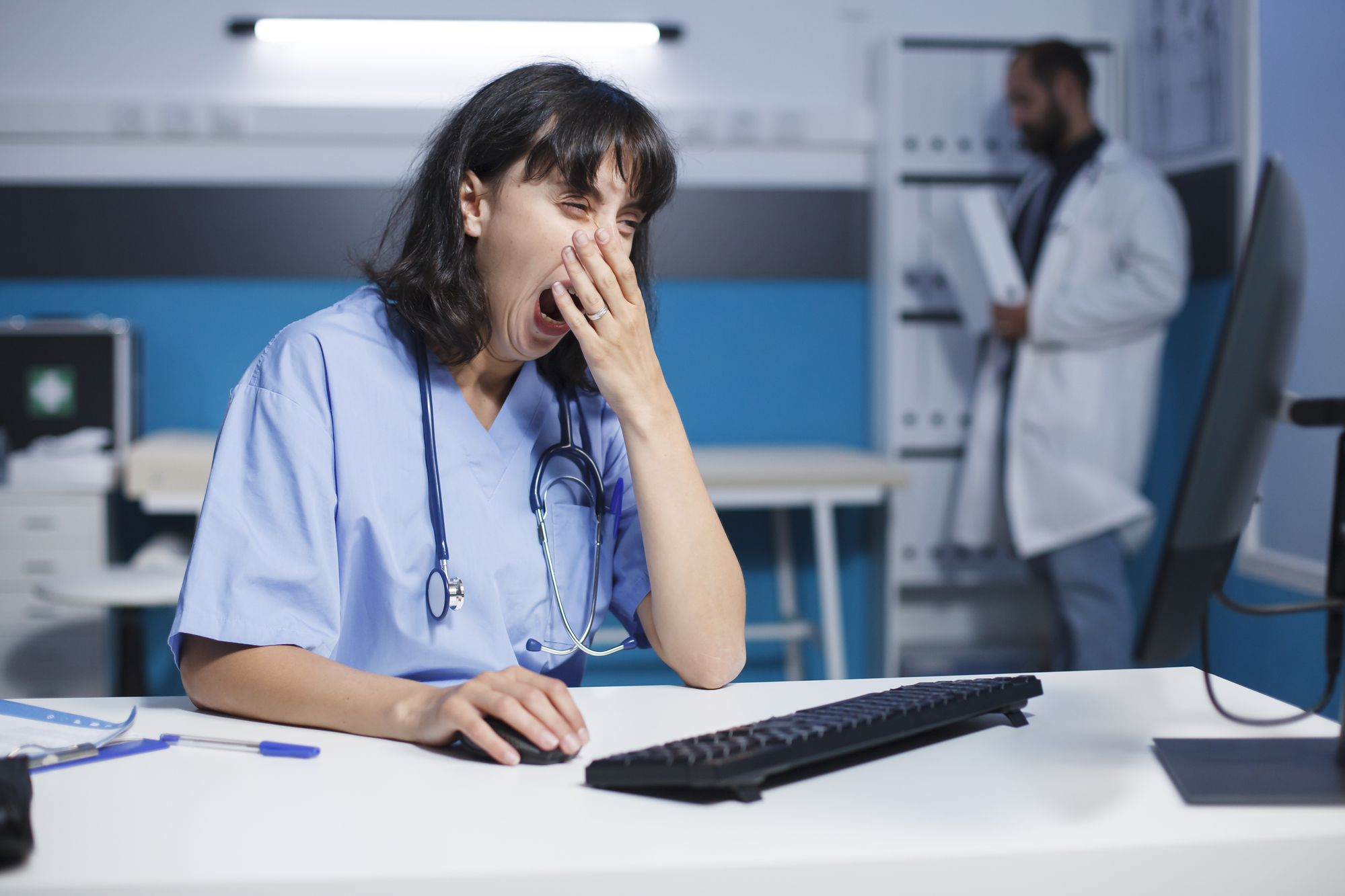 A tired healthcare worker, wearing blue scrubs and a stethoscope, sits at a desk in an office, yawning with one hand covering their mouth while the other is on a keyboard. In the background, another medical professional in a white coat is looking at a clipboard.